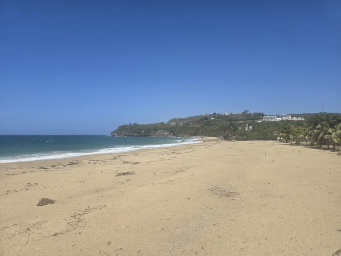 An empty sandy beach with gentle waves rolling onto the shore under a clear blue sky, with palm trees and buildings visible on a distant hillside.