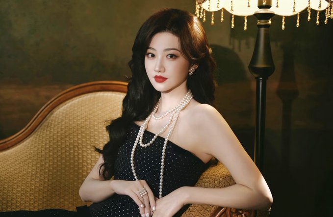 A young woman with long black hair sits elegantly on a vintage chaise lounge wearing a black polka dot dress and layered pearl necklaces, illuminated by a warm lamp in the background. The accompanying post references Justin Sun's lavish spending on private jet travel and luxury accommodations, with the image evoking the sophisticated, high-society lifestyle that such wealth can afford.