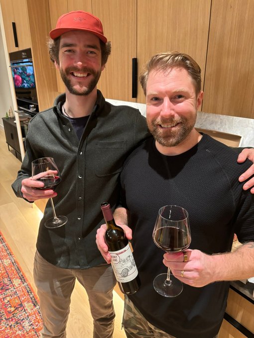 Two men smile together at an indoor gathering, one in a red cap holding a wine glass, the other holding a wine bottle and glass.