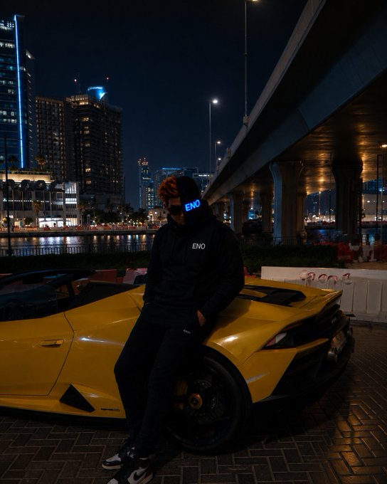A nighttime urban scene shows a person wearing an 'ENO' branded outfit leaning against a yellow sports car, with a city skyline and overpass illuminated in the background.