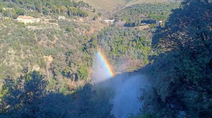 A scenic landscape view of a lush green canyon with a waterfall cascading down, creating a mist that forms a vibrant rainbow in the sunlight, with houses visible on the distant hillside.