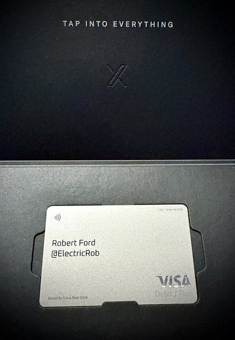 A metal Visa debit card issued by Cross River Bank with the name 'Robert Ford @ElectricRob' sits in a black wallet embossed with 'X' and the text 'TAP INTO EVERYTHING.'