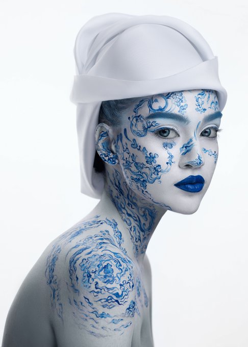A white-painted woman wearing a white head covering poses in profile, with intricate blue ornamental designs painted across her face, neck, and shoulder against a plain white background.