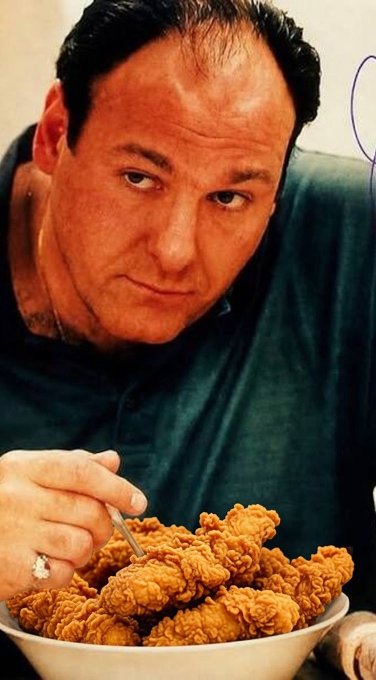 A man with slicked-back hair and a ring stares intensely at the camera while holding a fork over a large bowl of fried chicken.