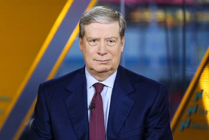 A male news anchor in a dark suit and red tie sits at a desk in a studio setting.