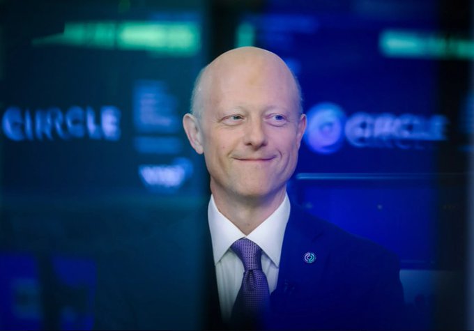 A bald man in a dark blue suit and tie smiling, standing in front of multiple screens displaying the 'CIRCLE' company logo with a blue background.