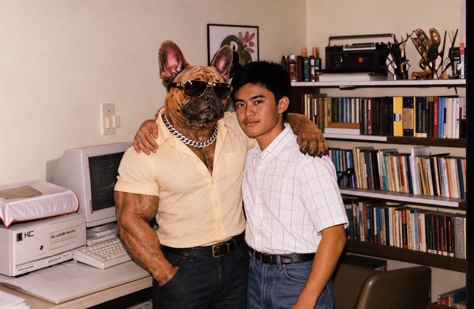 A vintage photograph showing a young man posing with a French Bulldog wearing sunglasses and a thick chain collar, in what appears to be a home office with a computer and bookshelves in the background.