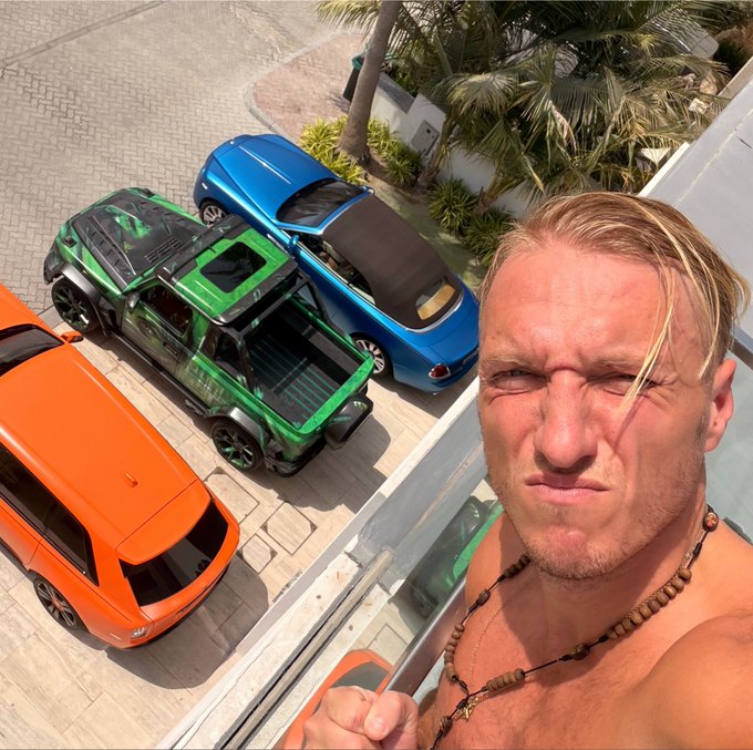A shirtless man with a serious expression takes a selfie from a balcony, with a bright orange SUV, a custom green pickup truck, and a blue sports car parked in the driveway below. The green pickup stands out as the unique 1/1 vehicle he's advertising for sale.