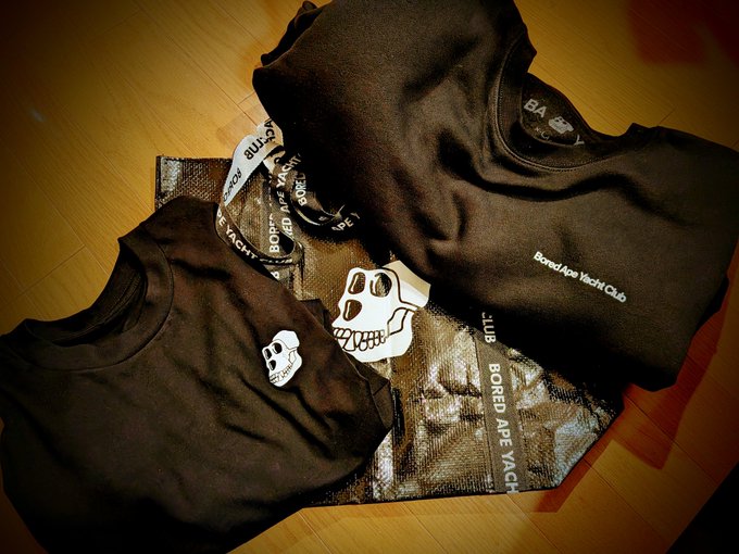The image features a brown t-shirt, possibly a sweatshirt or a hoodie, placed on top of two other shirts that are black and white. The shirts appear to be from the BAYC World Club, as indicated by the text "store.boredapeyachtclub.com". This suggests that these items might be merchandise related to the BAYC World Club, possibly for sale at a store or online marketplace.