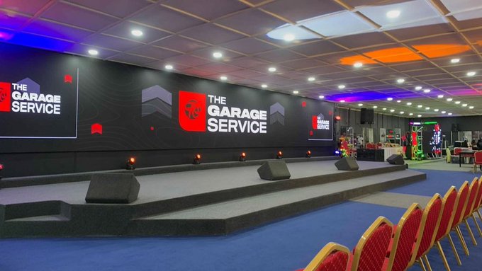 An empty event hall features a stage with large screens displaying "THE GARAGE SERVICE" logo, set up for a church rehearsal by an instrumentalist, while promotional branding for GalaxySwapLabs hints at an upcoming event or announcement.