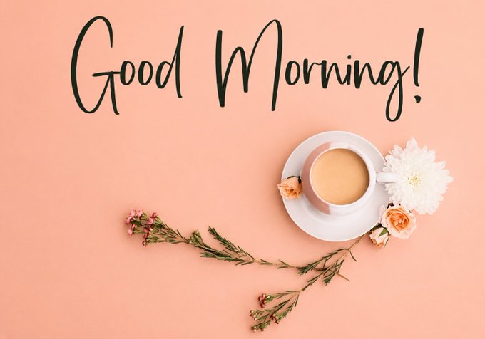 The image features a pink background with the words "Good morning!" written in black cursive font above a cup of coffee and flowers, which are placed on a table or countertop. The text "Good morning!" is positioned at the top center of the image, while the coffee cup and flowers are located towards the bottom right corner. The overall layout suggests that this image could be used as an eye-catching background for social media posts related to mornings, such as sharing a good morning message or promoting a new product like the coffee in the image.