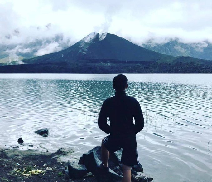 Silhouette of a person sitting on a rock by a calm lake, looking out at a misty volcanic mountain landscape.