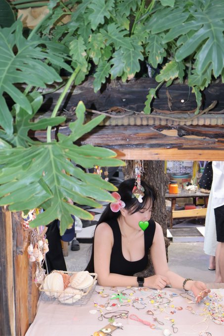 A young woman with a flower hair accessory and green leaf (likely a taste-testing mask) sits at a table displaying handmade jewelry and shell crafts in an outdoor tropical setting.