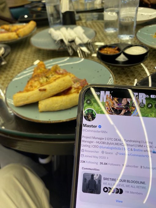 A smartphone displaying a Twitter/X profile for 'Master @Coinmaster100x' with bio 'Project Manager || OTC DEAL, Fundraising || Listing Manager' shown over a plate of pizza slices and breadsticks at a restaurant.