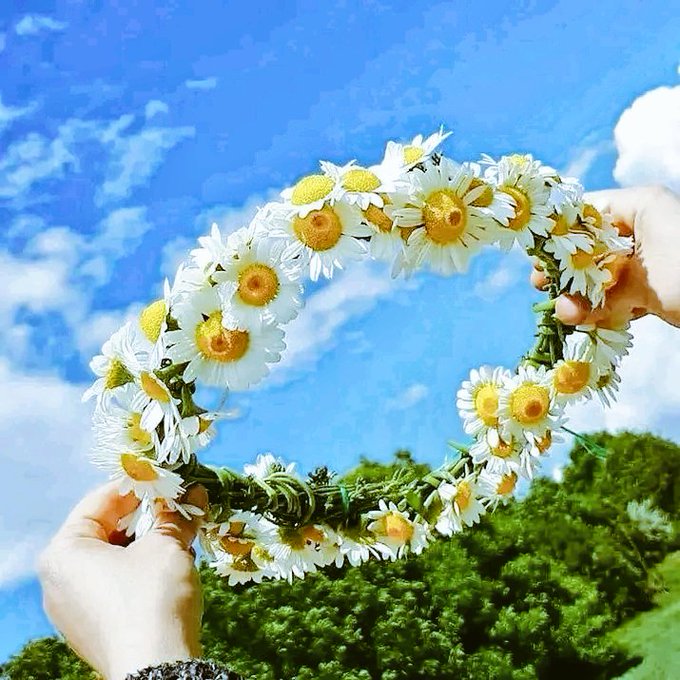 The image features two hands holding up a wreath made of white flowers against a blue sky with clouds. The wreath appears to be made from daisies, which are the primary color for this particular photo. The background includes trees, adding depth and context to the scene. The caption provided seems unrelated to the main subject matter or the image itself.