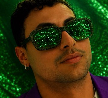A man wearing dark sunglasses stands against a sparkly green glitter backdrop, reflecting a glittering light in his lenses. His stylish, close-up portrait complements the post's playful, positive vibe about market rebounds, though it doesn't depict any specific Solana address or crypto-related content.