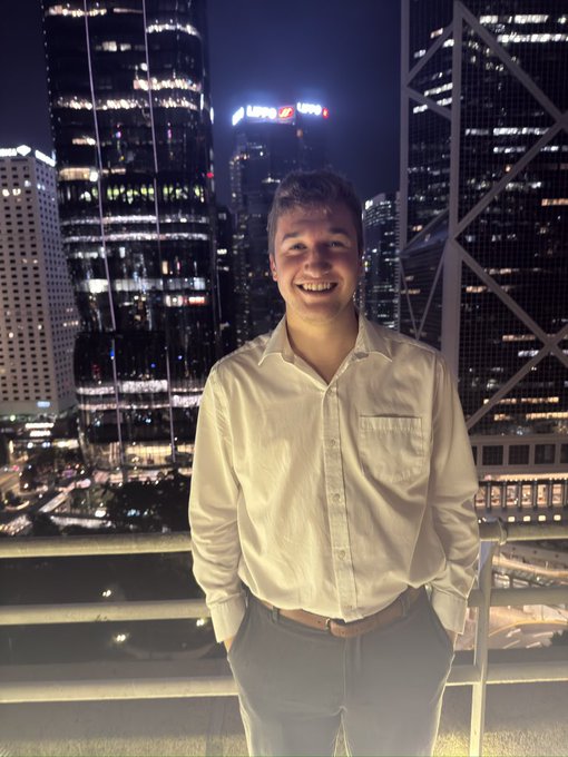 A smiling young person in a white button-up shirt and gray pants stands on a rooftop at night with an illuminated city skyline in the background.