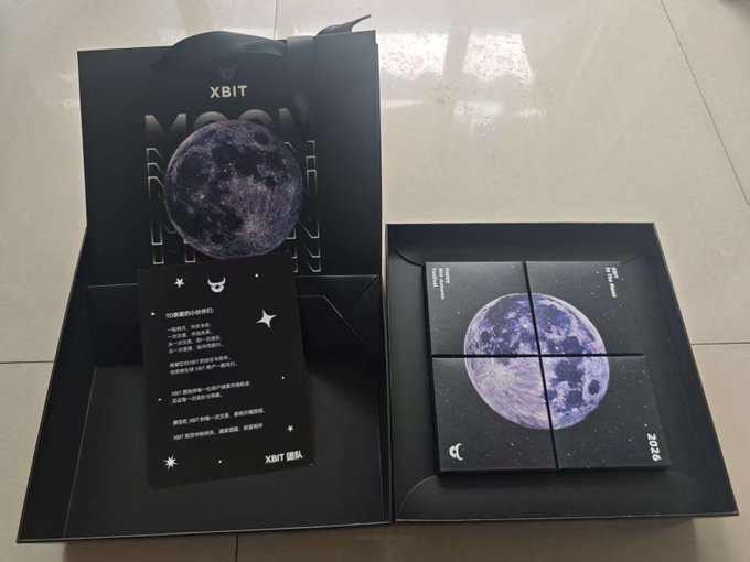 A black gift box and shopping bag from 'XBIT' display a moon-themed 2026 puzzle or calendar set with a card containing Chinese text, arranged on a tiled floor.