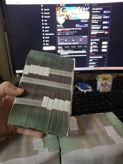 A hand holding a thick stack of cash, bundled with white paper bands, in front of a computer monitor. The monitor displays a YouTube live stream with a chat window on the right side.