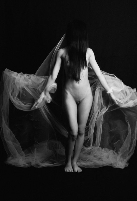 The image features a naked woman standing against a black background, with her arms outstretched as if she's about to take flight or reach for something. The photo appears to be in black and white, which adds an air of mystery and intrigue to the scene. The woman is positioned centrally within the frame, drawing attention to her form and expression.