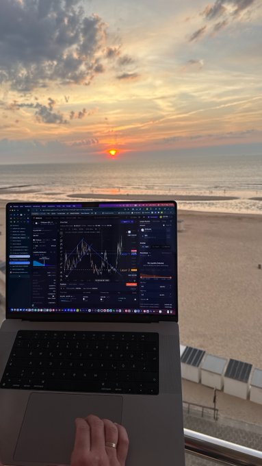A laptop displaying financial trading charts sits on a windowsill, with a hand resting on the trackpad and a scenic ocean sunset visible in the background.