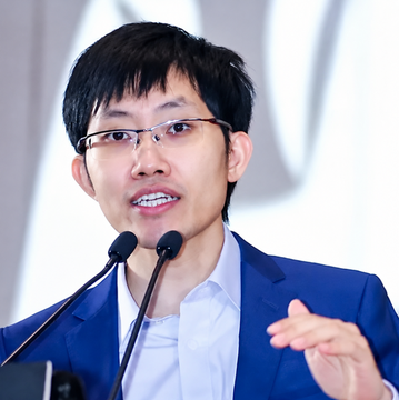 A man with glasses in a blue suit speaks at a podium with microphones, gesturing with his hand. The image likely depicts Liang Wenfeng, CEO of DeepSeek, referenced in the post about the suspended second fundraising round following leaked investor comments.