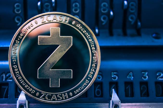 A close-up image of a physical Zcash (ZEC) cryptocurrency coin featuring its 'Z' logo and text reading 'decentralized', 'secured', and 'shielded' resting on a blue mechanical counter.