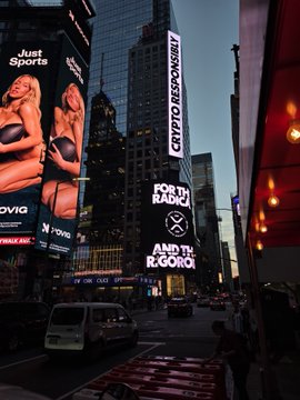 A nighttime cityscape, likely Times Square, features digital billboards displaying ads including 'Just Sports', 'CRYPTO RESPONSIBLY', and 'FOR THE AGENCY', with taxis on the road below.