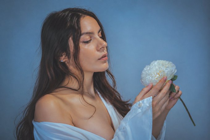 A woman with long dark hair, eyes closed, smelling a white peony flower against a blue background while wearing a light blue off-shoulder top.