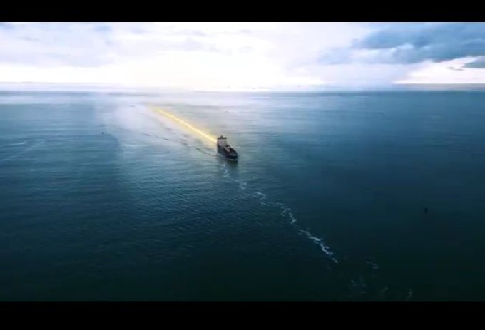 An aerial view of a cargo ship sailing across a vast calm ocean under a cloudy sky, leaving a white wake trailing behind it.