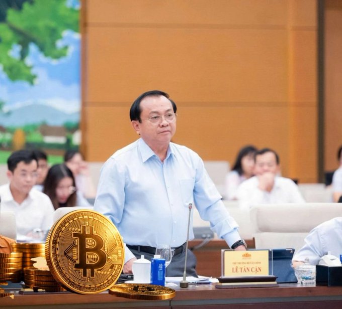 An official at a meeting with a nameplate reading 'LÊ TẤN CẬN', with a large Bitcoin coin and stacks of cryptocurrency coins placed on the desk.