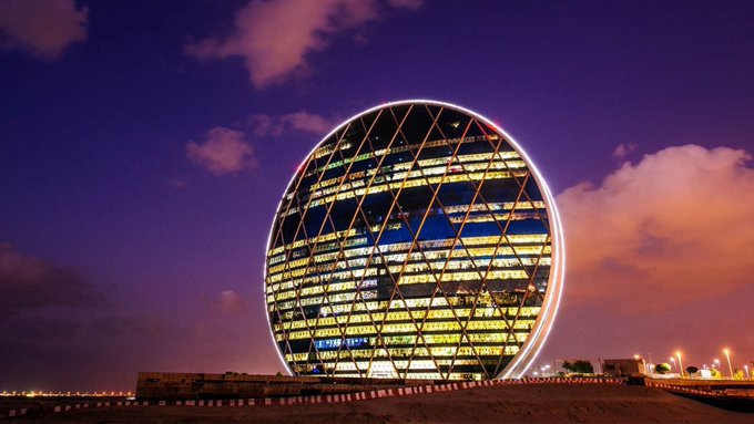 A circular, illuminated building with a geometric facade against a twilight sky.