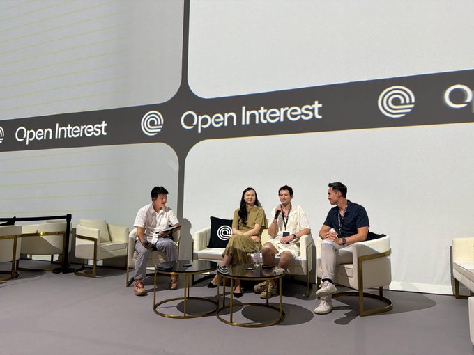 Four panelists sit in white chairs on a stage during an 'Open Interest' conference, with one person speaking into a microphone while others listen attentively.