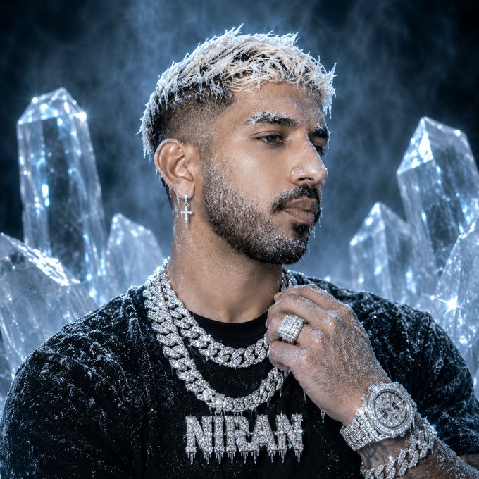 The image features a man wearing a black shirt with the name "Niran" written on it, along with a chain necklace and a watch. He has blonde hair and is looking off into the distance. The background of the photo consists of large crystals that appear to be made of ice or frozen water. The caption reads: "Solana."