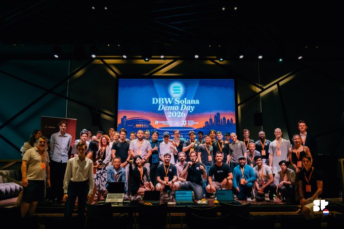 A group photo from the DBW Solana Demo Day 2026 event, showing attendees posing on stage in front of a large screen displaying the event branding.