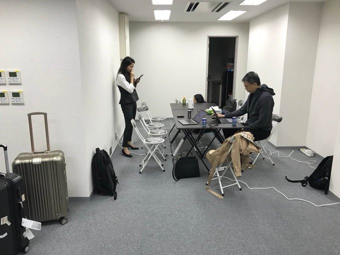An indoor office or meeting room with two people—one woman standing and looking at her phone, one man seated at a table working on a laptop—surrounded by luggage, backpacks, and folding chairs.