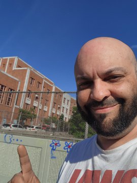 The image features a bald man with a beard, wearing a white t-shirt that says "I love you", standing next to a building under a blue sky. The man has his finger extended upwards, as if he's giving a thumbs up or making a gesture of approval. In the background, there are several cars parked on the street and a fence running along the edge of the property. The post includes text that reads "MATRIX TELLING ME NEW RAILS ON THE WAY @LOAN_Protocol @XPRNetwork".