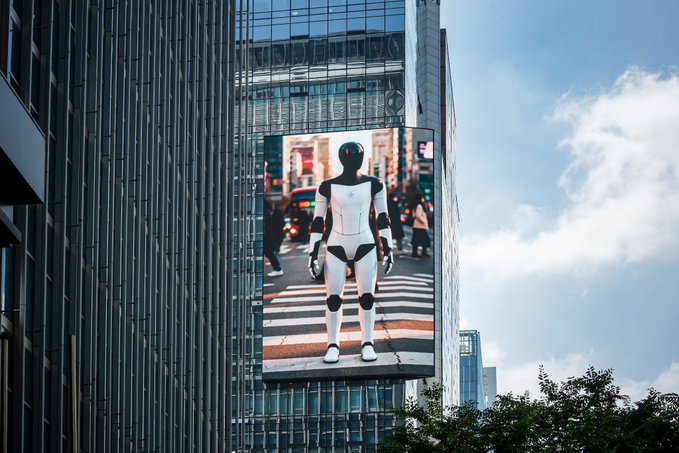 The image features a billboard with an advertisement for a robot, which appears to be a large screen TV mounted on top of a building. The billboard displays a picture of a robot standing at a crosswalk, surrounded by trees and buildings. This advertisement is promoting real adoption of robots in urban settings, as it showcases the integration of robots into everyday life.