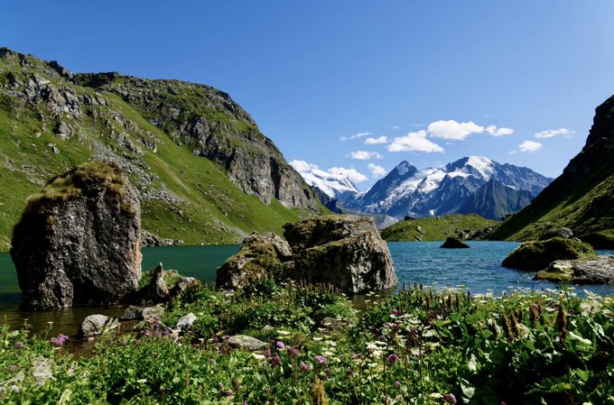 A scenic alpine landscape features a turquoise lake surrounded by lush green meadows, rocky outcrops, and snow-capped mountains under a blue sky.