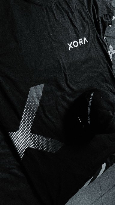 A close-up of black clothing items featuring the white text 'XORA' printed on the fabric, laid out on a dark surface.