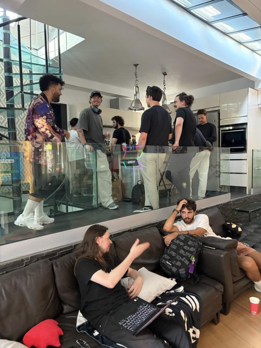 A casual gathering in a modern home with a group of people conversing in an elevated kitchen area while others lounge on a brown leather sofa in the foreground.