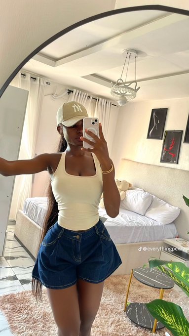 A woman takes a mirror selfie in a bright bedroom, wearing a beige cap with "NY" embroidery, a cream tank top, and a denim mini skirt. The TikTok watermark "@everythingpea..." is visible, fitting the post's casual "Gm" morning greeting to the @0xMiden community as she starts her day focused on consistency and growth.
