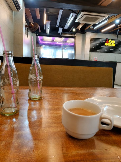 A white coffee cup and an empty glass bottle with a pink and white striped straw sit on a wooden table in a casual dining space with an air conditioning unit and 'FOR CORAM' sign in the background.