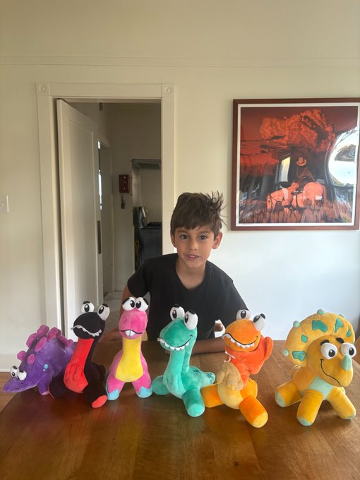 A young boy with messy hair sits at a wooden table displaying a handcrafted row of colorful clay dinosaur figures in purple, red, yellow, teal, orange, and yellow-green.