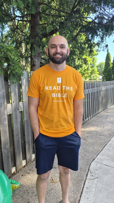 A smiling, bald man with a beard standing on a sidewalk next to a wooden fence, wearing a yellow t-shirt that says 'READ THE BIBLE' with a Bible icon.