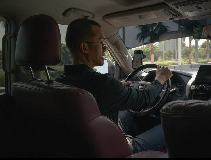 A man wearing glasses and a dark jacket drives a car, gripping the steering wheel with a phone mounted on the dashboard and palm trees visible through the windshield.