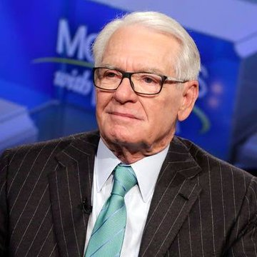 A photograph of an elderly man with white hair and glasses, wearing a dark pinstripe suit with a green tie, speaking on a panel with partial 'Me' text visible in the background.