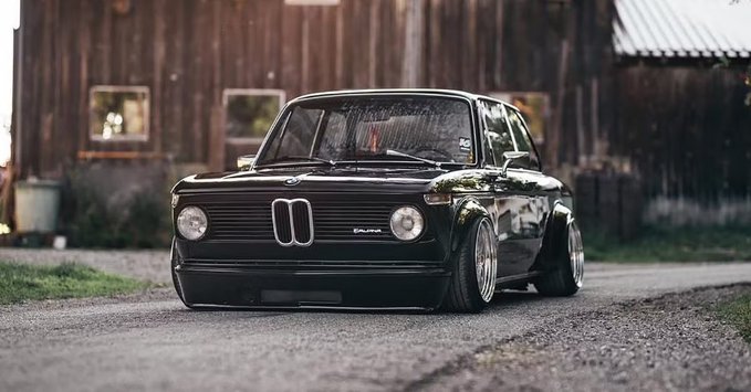 A black classic BMW 2002 Alpina is parked on a gravel road in front of a rustic wooden building.