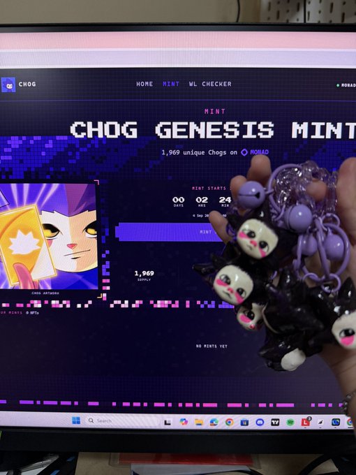 A hand holding several black and white cat-shaped keychains in front of a computer monitor displaying the 'CHOG GENESIS MINT' website on Ronad, showing '1,969 unique Chogs' with a countdown timer.