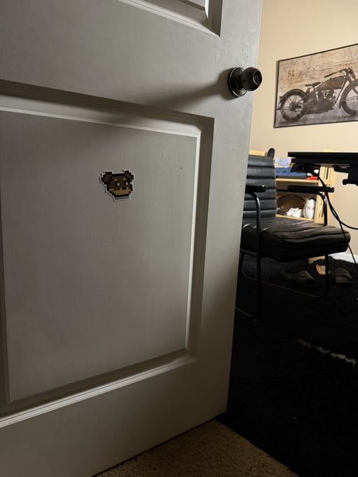 A partially open door with a Space Invaders sticker on it reveals a room with a leather chair and a motorcycle poster featuring the number "66," creating a personal, reflective atmosphere that complements the post's contemplative mood about community and disconnection in crypto.